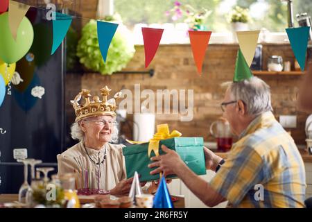Seniorenpaar feiert zusammen, ältere Geburtstagsfrau erhält Geburtstagsgeschenk von ihrem Mann, der zu Hause am Esstisch sitzt. Celebarati Stockfoto