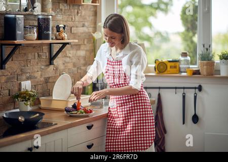 Fröhliche junge Hausfrau, die in einer modernen Küche Gemüsesalat aus vegetarischen Scheiben auf einem Teller macht. Gesundes Essen, Zuhause, Lifestyle-Konzept. Stockfoto