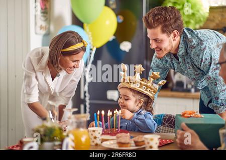 Ein Geburtstagskind, das als Geburtstagskrone goldene aufblasbare Kronen trägt, am Tisch sitzt, mit Eltern in der Nähe eines Großvaters, jubelt sie an. Zuhause, Familie, Cel Stockfoto