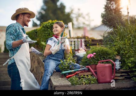 Ein junger Vater mit einer Tochter, die im Blumengarten beim Haus zusammenarbeitet, ein Kind hält einen kleinen Gartenrechen bereit, Blumen zu Pflanzen. Nach Hause, Familie Stockfoto