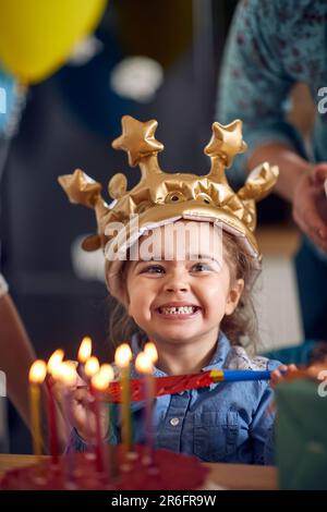 Ein bezauberndes kleines Geburtstagskind, das aufblasbare goldene Kronen trägt, vor dem Kuchen sitzt, mit brennenden Kerzen, lächelt und sich fröhlich fühlt. Geburtstag, Feier Stockfoto