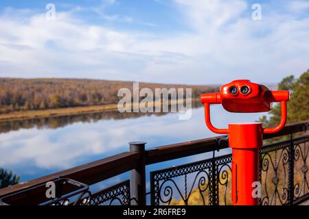 Öffentliche stationäre Ferngläser am Flussufer im Sommer oder Herbst, um die Natur zu erkunden, münzbetriebene rote Ferngläser aus Metall. Stockfoto