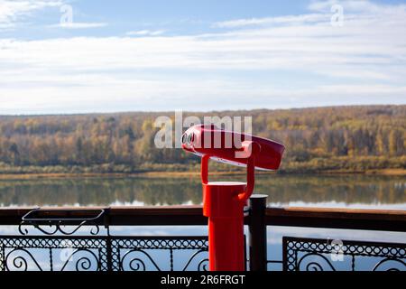 Öffentliche stationäre Ferngläser am Flussufer im Sommer oder Herbst, um die Natur zu erkunden, münzbetriebene rote Ferngläser aus Metall. Stockfoto