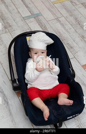 Ein asiatisches Kind mit Köchmütze und Schürze hat mit seiner mutter Weihnachtsginger-Kekse gekocht und ruht sich jetzt in seinem Kinderwagen aus und isst aus einer Flasche Stockfoto