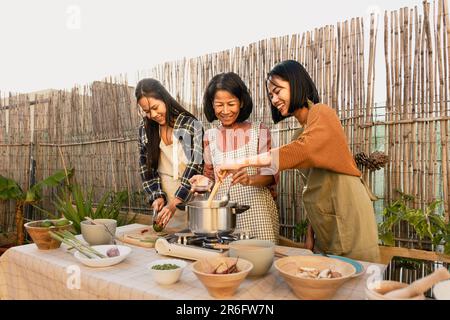 Südostasiatische Mutter und ihre Töchter haben Spaß dabei, gemeinsam auf der Terrasse des Hauses ein Rezept zuzubereiten Stockfoto