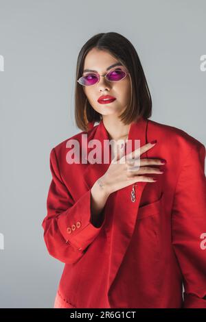 Moderne Mode, junge brünette Frau mit kurzen Haaren in pinkfarbener Sonnenbrille und rotem Anzug auf grauem Hintergrund, Generation z, trendiges Outfit, fas Stockfoto