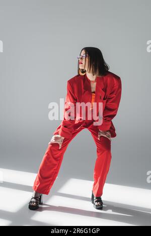 Business-Mode, junge brünette Frau mit kurzen Haaren in pinkfarbener Sonnenbrille und rotem Anzug auf grauem Hintergrund, Generation z, trendiges Outfit, Mode Stockfoto