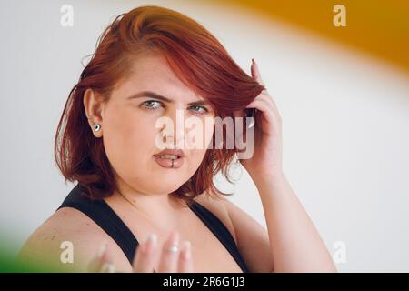 Nahaufnahme einer jungen, übergroßen argentinischen Latina-Frau mit grünen Augen und rotem Haar, die in die Kamera schaut und farbige Ohrringe um sie herum trägt. Stockfoto