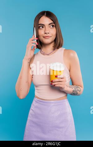 Zum Kaffee, glückliche und tätowierte junge Frau mit kurzen Haaren und Nasenlöchern, die einen Pappbecher in der Hand hält und auf einem Smartphone auf blauem Hintergrund spricht, Generat Stockfoto