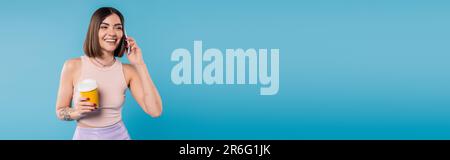 Kaffee zum Mitnehmen, fröhliche und tätowierte junge Frau mit kurzen Haaren und Nasenlöchern, die einen Pappbecher hält und auf einem Smartphone spricht, auf blauem Hintergrund, Genera Stockfoto