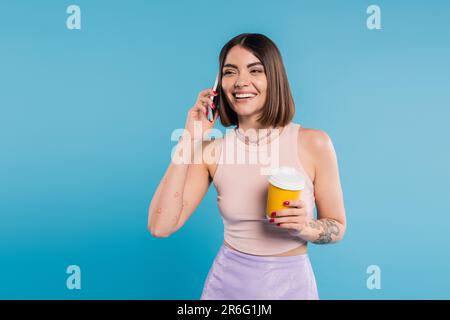 Zum Kaffee, fröhliche und tätowierte junge Frau mit kurzen Haaren und Nasenlöchern, die einen Pappbecher hält und auf einem Smartphone spricht, auf blauem Hintergrund, Genera Stockfoto