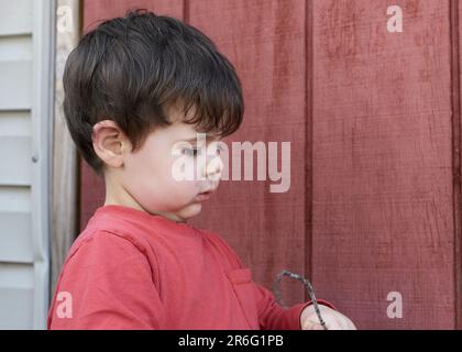 Süßer kleiner Junge, der mit Zweigen im Garten bei der Garage spielt Stockfoto