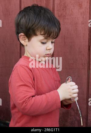 Süßer kleiner Junge, der mit Zweigen im Garten bei der Garage spielt Stockfoto