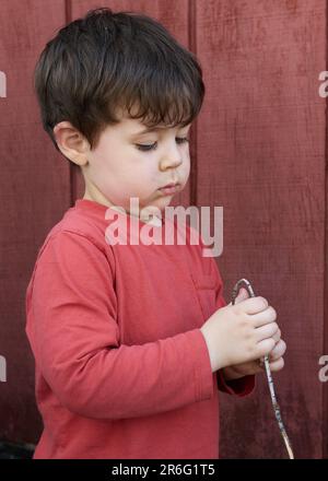 Süßer kleiner Junge, der mit Zweigen im Garten bei der Garage spielt Stockfoto