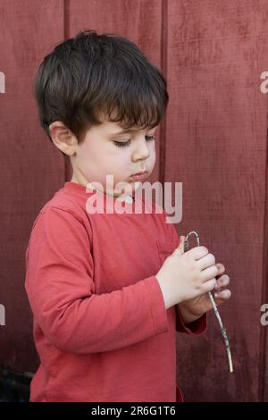 Süßer kleiner Junge, der mit Zweigen im Garten bei der Garage spielt Stockfoto