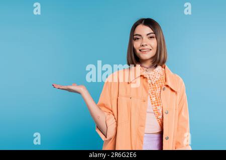 Glückliche junge Frau mit kurzen Haaren, Piercing in der Nase und Tätowierungen, die etwas in der Kamera auf blauem Hintergrund zeigt, orangefarbenes Hemd, Summer ou Stockfoto