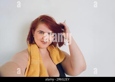 Selfie-Porträt einer jungen, übergroßen, argentinischen Latina-Frau, mit hellen Augen und roten Haaren, lächelnd und schauend in die Kamera, in schwarzer Sportbekleidung Stockfoto