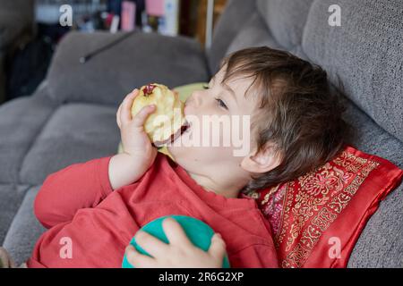 Ausdrucksstarker Junge, der einen Apfel isst und sich zu Hause auf der Couch ausruht Stockfoto