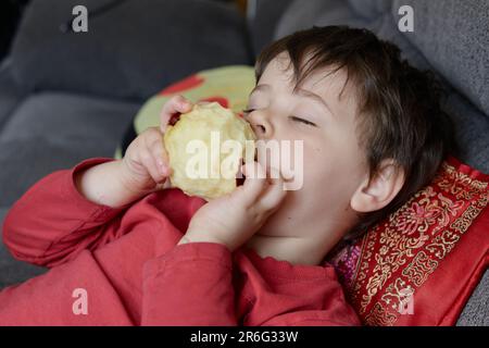 Ausdrucksstarker Junge, der einen Apfel isst und sich zu Hause auf der Couch ausruht Stockfoto