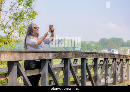 Reife Frau, die auf einer Holzbrücke steht und Fotos mit ihrem Handy, Bäumen und Dorf im nebligen Hintergrund macht, sonniger Frühlingstag in holländischer Natur Stockfoto