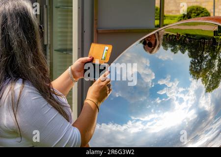Reife Touristin, die Fotos in einem runden Spiegel macht, einen Teil ihres Gesichts, blauen Himmel mit weißen Wolken und grünen Bäumen, die sich in der Spiegeloberfläche spiegeln Stockfoto