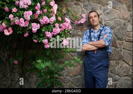 Lächelnder, gut aussehender Mann, Blumenhändler in blauer Arbeitskleidung, schaut in die Kamera, steht im Garten einer Villa Stockfoto