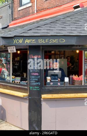 Toronto, Kanada - 4. Juni 2023: Kensington Market. Eine junge Frau, die als Kassiererin in einer Cafeteria arbeitet. Die Außenfassade des Ladens trägt die Aufschrift „Yo Stockfoto