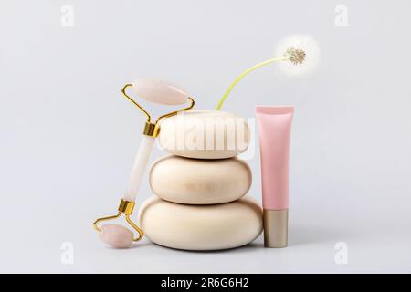 Anti-Age-Gesichtsmassage für natürliches Heben und zu Hause. Gesichtswalze aus Rosenquarzglas und Kosmetikflaschenbehälter auf Holzsteinen mit Blumendandelio Stockfoto