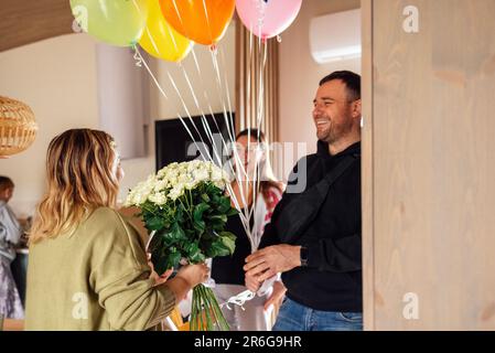 Das junge Paar gratuliert der blonden Frau zum Geburtstag. Eine attraktive Frau in Brille gibt einen Strauß weißer Blumen und küsst das Geburtstagskind. Die Hände Stockfoto