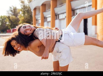 Zwei ethnische weibliche Teenager, die sich umarmen und Spaß miteinander haben. Fröhliche Freunde in lässigen Klamotten, die herumalbern und auf der Straße lachen. Süßer Junge Stockfoto