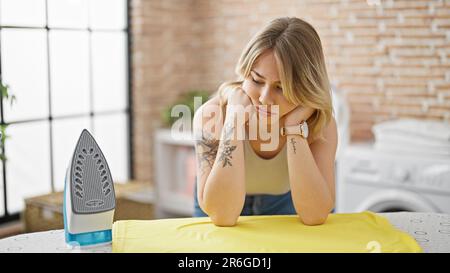 Junge blonde Frau, die sich auf den Bügeltisch lehnt, mit traurigem Gesichtsausdruck in der Wäscherei Stockfoto