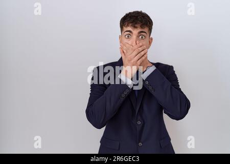 Junger hispanischer Geschäftsmann, der Anzug und Krawatte trägt, hat einen schockierten Mund mit falschen Händen bedeckt. Geheimes Konzept. Stockfoto