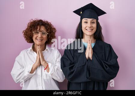 Hispanische Mutter und Tochter tragen Abschlussmütze und Zeremonie-Morgenmantel, beten mit den Händen zusammen und bitten um Vergebung, lächelnd selbstbewusst. Stockfoto