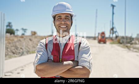 Junger hispanischer Baumeister, der selbstbewusst lächelt, mit gedrückten Armen auf der Straße Stockfoto