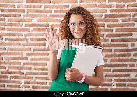 Junge weiße Frau in der Hand eines Art Notizbuchs, die auf Hallo verzichtet, fröhlich und lächelnd, freundliche Begrüßungsgeste Stockfoto