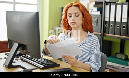 Junge rothaarige Geschäftsangestellte, die im Büro einen Briefumschlag zum Öffnen des Computers benutzt Stockfoto