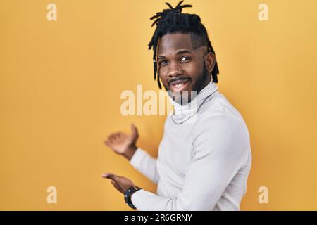 Ein afrikanischer Mann mit Dreadlocks trägt einen Rollkragenpullover auf gelbem Hintergrund und lädt dazu ein, mit offener Hand zu lächeln Stockfoto