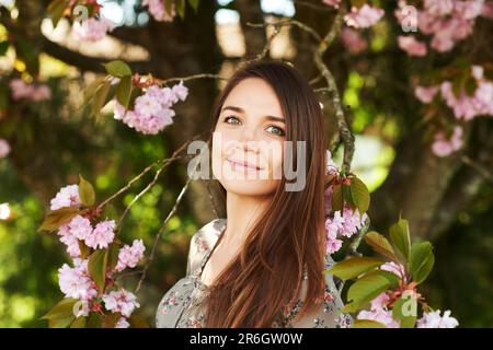 Freiluft-Frühlingsporträt einer hübschen jungen Frau mit braunem Haar, die unter einem rosa blühenden Baum posiert Stockfoto