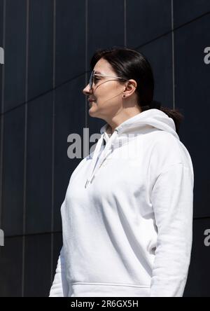 Portrait Pretty Mature Brunette Woman in White Hoodie und Sonnenbrille Looks Away, Dark Background Wall. Sportlich Selbstbewusst 40 Yo Wunderschön Weiß Stockfoto