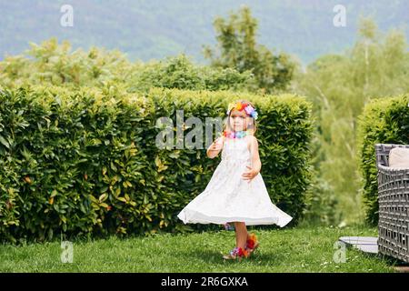 Außenporträt eines niedlichen kleinen Mädchens, das im Park spielt, weißes Kleid trägt und farbenfrohe Blumen trägt Stockfoto