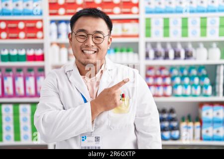 Chinesischer junger Mann, der in einer Apotheke arbeitet, fröhlich mit einem Lächeln auf dem Gesicht, der mit Hand und Finger zur Seite zeigt, mit fröhlichem und natürlichem Ausdruck Stockfoto