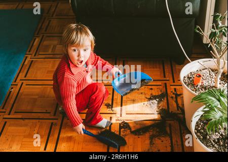 Sehr unordentliches Baby mit lustigem Gesichtsausdruck, das Pflanzenerde vom Boden entfernt Stockfoto