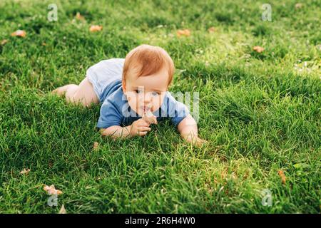 Süßer rothaariger Junge, der im Sommerpark auf frischem, grünem Gras krabbelt und trockenes Blatt isst Stockfoto