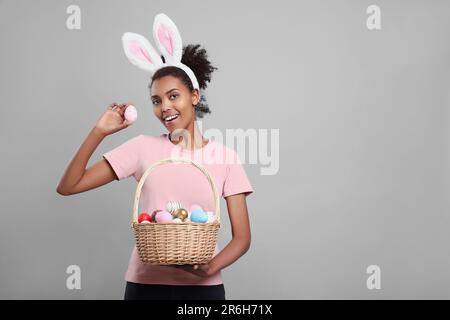 Glückliche afroamerikanische Frau mit Hasenohren Stirnband mit Korb aus Korb mit Ostereiern auf grauem Hintergrund, Platz für Text Stockfoto