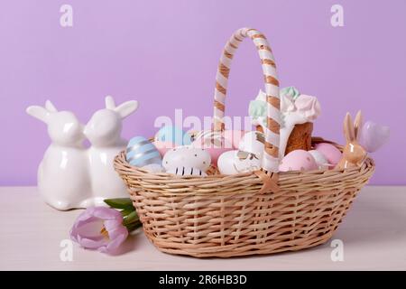 Osterkorb mit bemalten Eiern, Kuchen, Blumen und Kaninchen auf einem weißen Holztisch Stockfoto