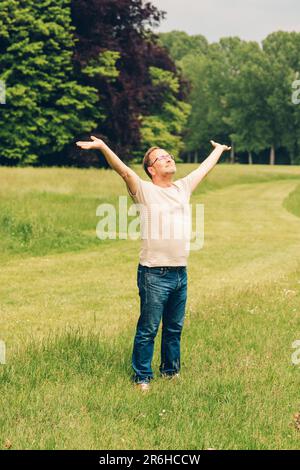 Glücklicher Mann mittleren Alters, der einen schönen Tag im Sommerpark genießt und die Arme weit offen hält Stockfoto