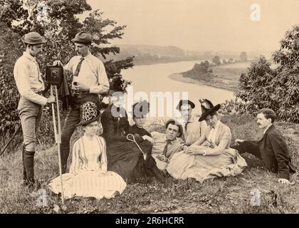 1890S FAMILIENGRUPPE, DIE AUF DEM HÜGEL MIT BLICK AUF DEN FLUSS SITZT, WÄHREND FOTOGRAF UND ASSISTENT DIE KAMERA FÜR DAS BILD VORBEREITEN - O2392 SPL001 UHR HARS DAMEN NACHDENKLICHE MÄNNER MITTELALTER 1800S SCHWARZWEISS BLICKKONTAKT MITTLERE ALTERS FRAU HOCHWINKEL ABENTEUER FREIZEIT BEREITEN AUFREGENDE FREIZEITAKTIVITÄTEN VOR ESCAPE-FRIENDLY NEUN STILVOLLE, AUFRICHTIGE 9 FOKUSSIERTE HÜGEL INTENSIV MITTELERWACHSENER MANN MITTELERWACHSENER FRAU MIT BLICK AUF ENTSPANNUNG ZUSAMMEN JUNGER ERWACHSENER MANN, JUNGE ERWACHSENE FRAU, SCHWARZ-WEISS, VORSICHTIG, KAUKASISCHE ETHNISCHE HERKUNFT, ERNSTE ABSICHT ALTMODISCH Stockfoto