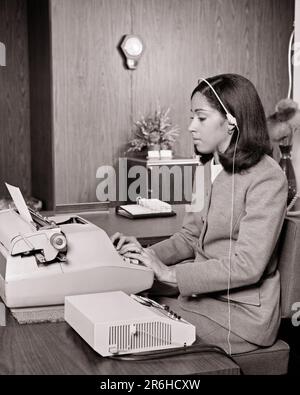 1970S AFROAMERIKANISCHE FRAU IM BÜRO, DIE AUF EINER ELEKTRISCHEN SCHREIBMASCHINE SCHREIBT UND DIKTAT VOM DIKTAT MIT HEADSET HÖRT - O3035 HAR001 HARS HALBLANGE DAMEN VERTRAUEN SCHWARZWEISS-HEADSET DIKTIERGERÄT FÄHIGKEITEN FÄHIGKEITEN SCHREIBEN AFROAMERIKANISCHE AFROAMERIKANISCHE SCHWARZE ETHNISCHE BÜROANGESTELLTE BERUFE GAL FRIDAY ADMINISTRATOR-LISTE SEKRETÄRINNEN STILVOLLE UNTERSTÜTZUNG AMANUENSIS-DIKTAT AUFGEZEICHNET JUNGE ERWACHSENE FRAU SCHWARZ-WEISS KLERIKER HAR001 ALTMODISCHE SCHREIBKRAFT AFRIKANER AMERIKANER Stockfoto