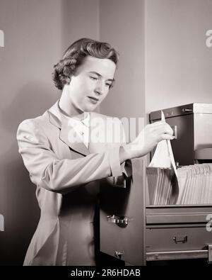 1950S BRÜNETTE FRAU, DIE IN EINEM AKTENSCHRANK ARBEITET, ENTFERNT ODER LEGT PAPIERKRAM AB - O658 HAR001 HALBLANGE DAMEN MIT HARS, DIE AUFMERKSAM SIND CARING CONFIDENCE SCHWARZWEISS-DATEIEN ENTFERNEN FRISUREN BÜROANGESTELLTE ARBEIT BÜROANGESTELLTE BESCHÄFTIGUNGEN GAL FRIDAY ADMINISTRATOR SEKRETÄRE EHRLICHE MITARBEITER ODER FEIERLICHE AMANUENSIS FOKUSSIERTE, INTENSIVE JUNGE ERWACHSENE FRAU SCHWARZ-WEISS VORSICHTIG KAUKASISCHE ETHNISCHE ZUGEHÖRIGKEIT BÜROANGESTELLTER ERNSTHAFT DATEI HAR001 INTENT SIE ARBEITEN ALTMODISCH Stockfoto