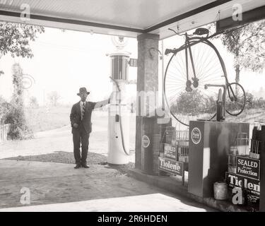 1940S-MANN-BESITZER, DER SICH AN DER TANKSTELLE MIT EINER TANKSTELLE MIT EINEM ANTIKEN PENNY-FARTHING-BIKE UND EINEM MUSKETEN-SCHLANGENHÜGEL IN OHIO BEDIENT USA - Q43998 CPC001 HARS KOPIERRAUM VOLLE LÄNGE PERSONEN UNITED STATES OF AMERICA MÄNNER KRAFTSTOFF VERTRAUEN FAHRRÄDER TRANSPORT B&W MITTLEREN ALTERS BIKES NORDAMERIKA MANN MITTLEREN ALTERS NORDAMERIKANISCHE ALTERS ÄLTERE TANKSTELLEN TANKSTELLEN TANKSTELLEN TANKSTELLEN TANKSTELLEN TANKSTELLEN ANTIQUITÄTEN BERUFE ÄLTERE BENZINBESITZER OHIO, GEWÖHNLICHER PENNY-FARLING, BENZIN, SCHWARZ-WEISS, WEISS, WEISS, WEISSE ETHNISCHE HERKUNFT, ALTMODISCH Stockfoto